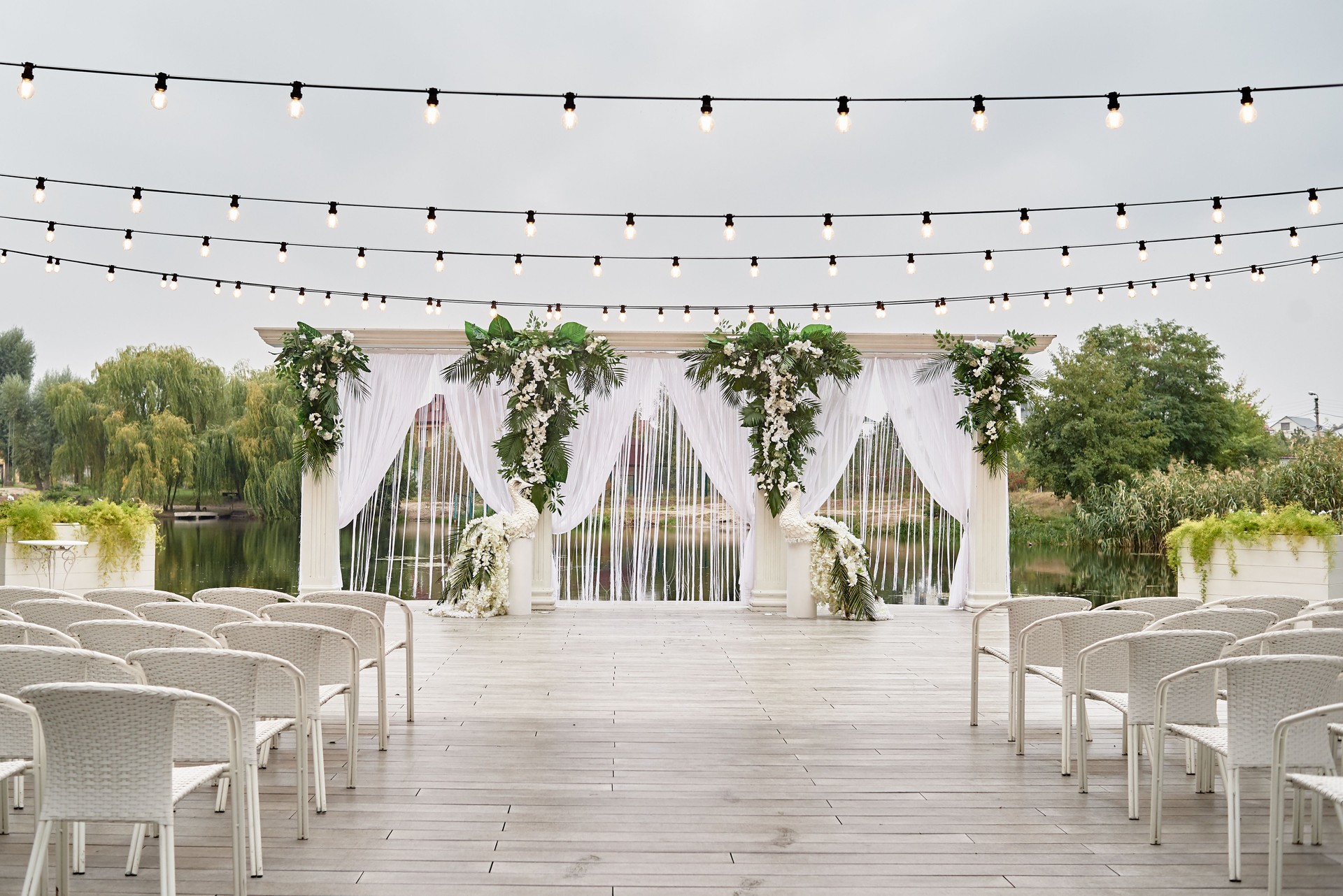 Place for wedding ceremony with wedding arch decorated with palm leaves, orchid flowers and floral peacocks, bulbs garland and white chairs outdoors, copy space