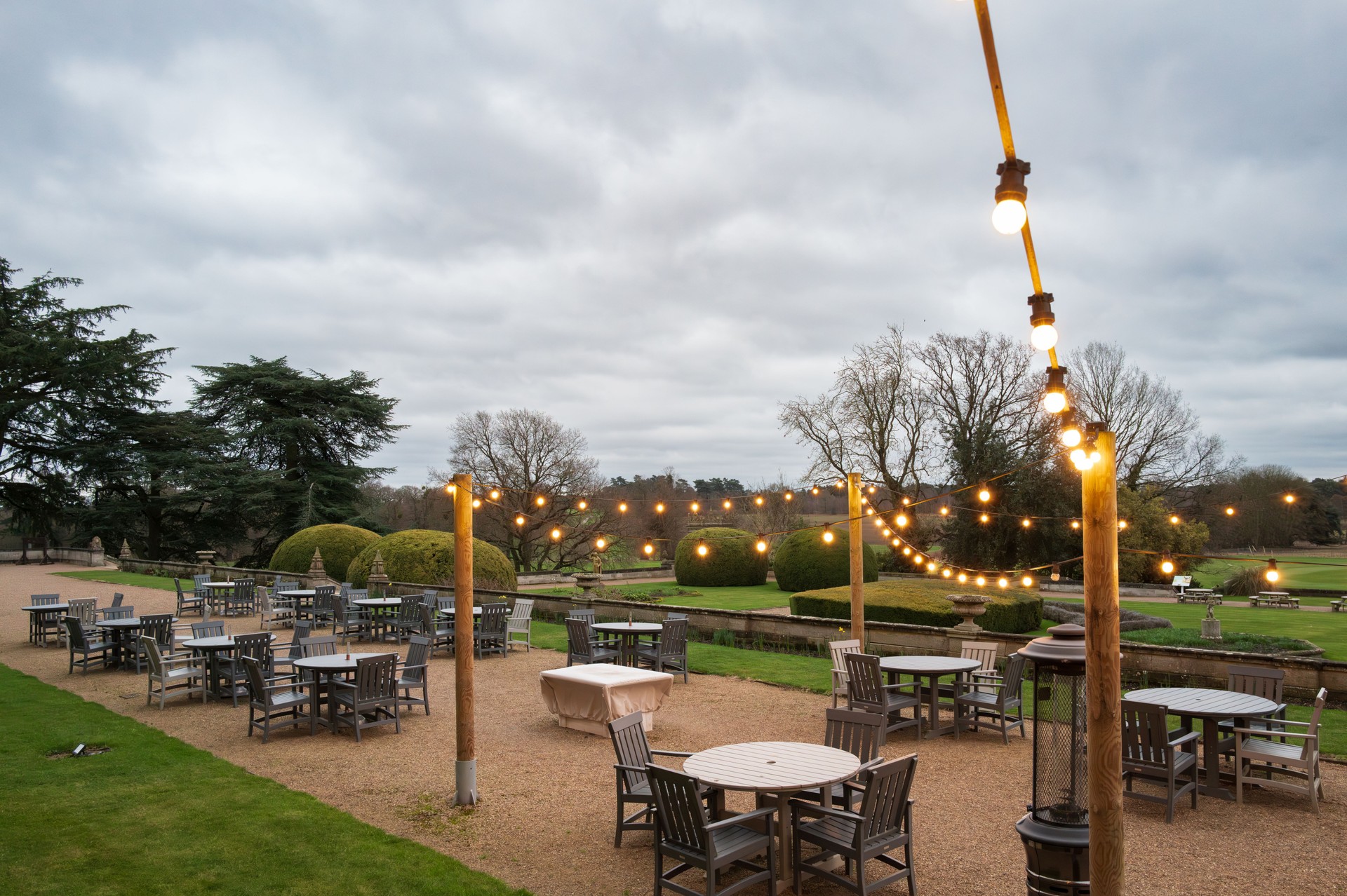 Warm string lights seen strewn above a large outside dining area at a country house and statley home in rural mid England.